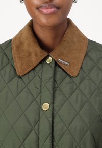 Επενδυμένος ελαιοπράσινος μπουφάν με καφέ κοτλέ γιακά, χρυσά κουμπιά και ασημένια ετικέτα με λογότυπο κοντά στον γιακά.