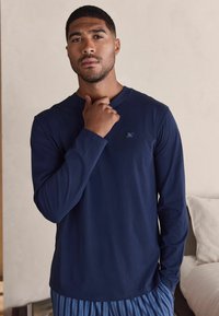 Jūras spēku garu piedurknēm kreklis, kas izgatavots no mīksta auduma, ar apaļu kakla izgriezumu un mazu izšūtu logotipu uz krūtīm. Saskaņots ar svītrainām biksēm.