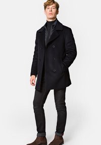 Czarny wełniany płaszcz typu peacoat z podwójnym zapięciem, dużym kołnierzem i przednimi kieszeniami. Połączony z czarnymi zniszczonymi dżinsami i brązowymi zamszowymi botkami.