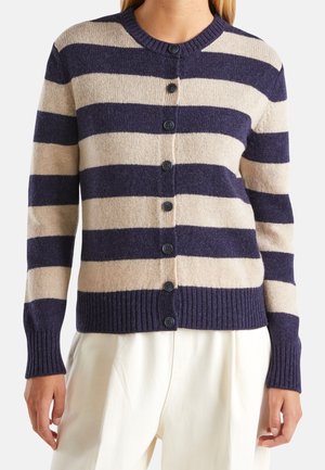 Žena nosící námořnicky modrý a béžový pruhovaný zapínací svetr s dlouhými rukávy a bílé kalhoty.
