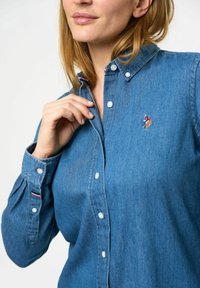 Denim overhemd in het blauw, met een kraag, witte knopen en een klein geborduurd logo. Opgestikte mouwen met gestreept detail.