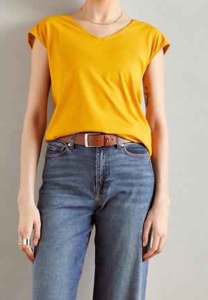 Persoon draagt een geel kortemouw-t-shirt ingestopt in een blauwe spijkerbroek met een bruine riem, staand tegen een neutrale achtergrond.