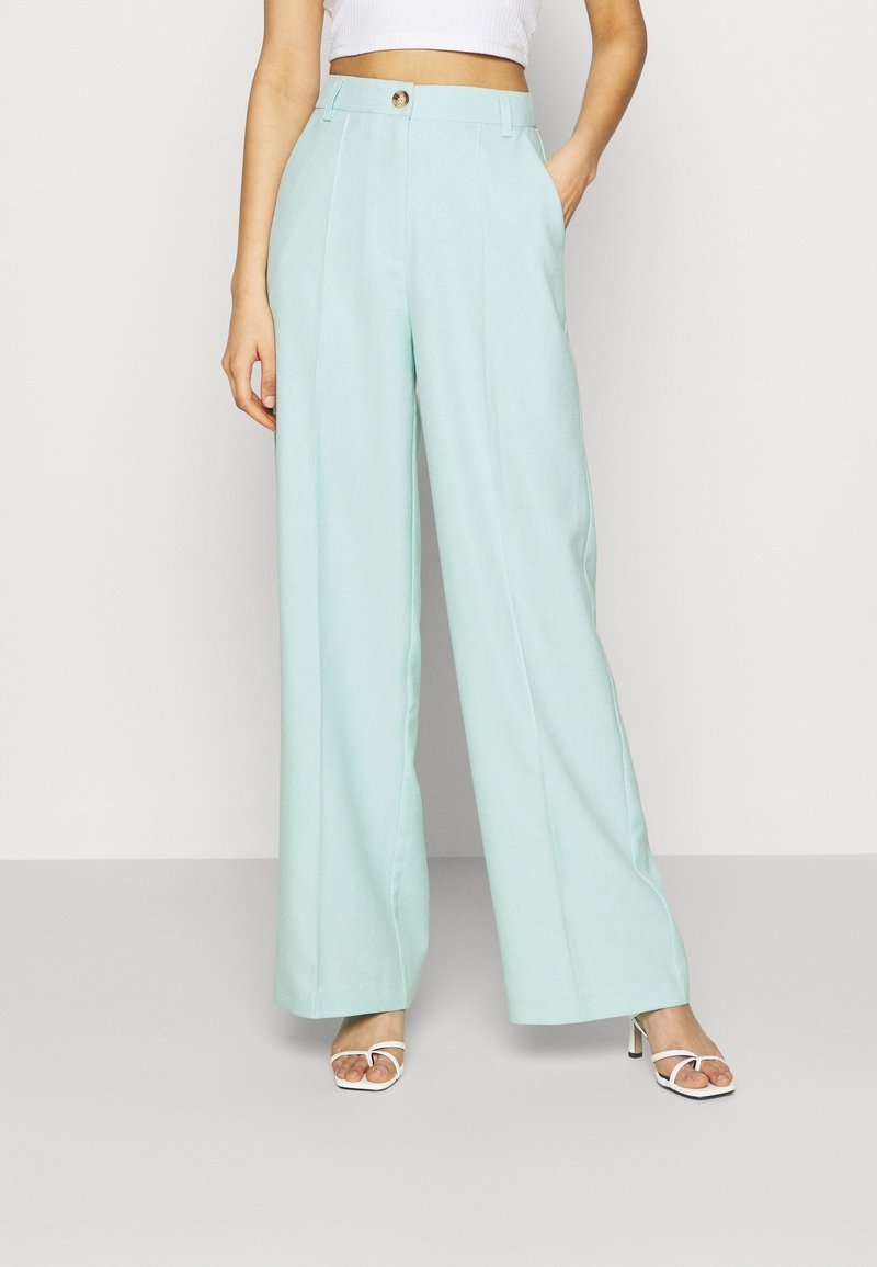 Personne portant un pantalon bleu clair taille haute, à jambes larges, associé à des sandales blanches à talons hauts et à lanières, et un top blanc court.