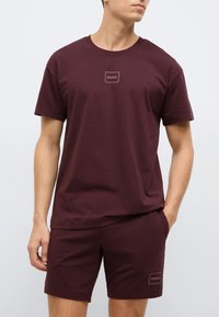 Viininpunainen puuvillasekoitetuista materiaaleista valmistettu t-paita ja shortsit, joissa molemmissa vasemmassa rinnassa ja shortseissa on pieni samalla värillä oleva logopatch.