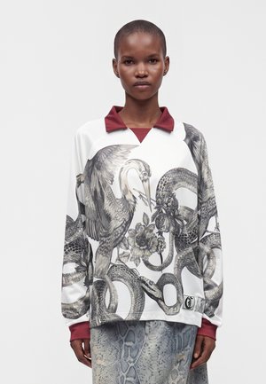 Persona che indossa una camicia a maniche lunghe con un motivo di uccello, serpente e fiori, colletto e polsini color bordeaux, abbinata a pantaloni grigi fantasia.