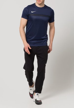 Man loopt, draagt een donkerblauw Nike T-shirt, zwarte Nike joggingbroek en witte Nike sneakers tegen een effen achtergrond.