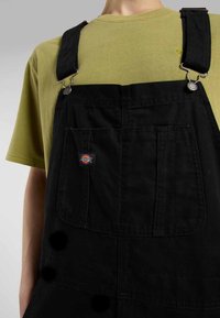 Salopette noire avec des bretelles réglables et du matériel en métal argenté, comportant des poches avant et un patch logo. T-shirt vert olive en dessous.