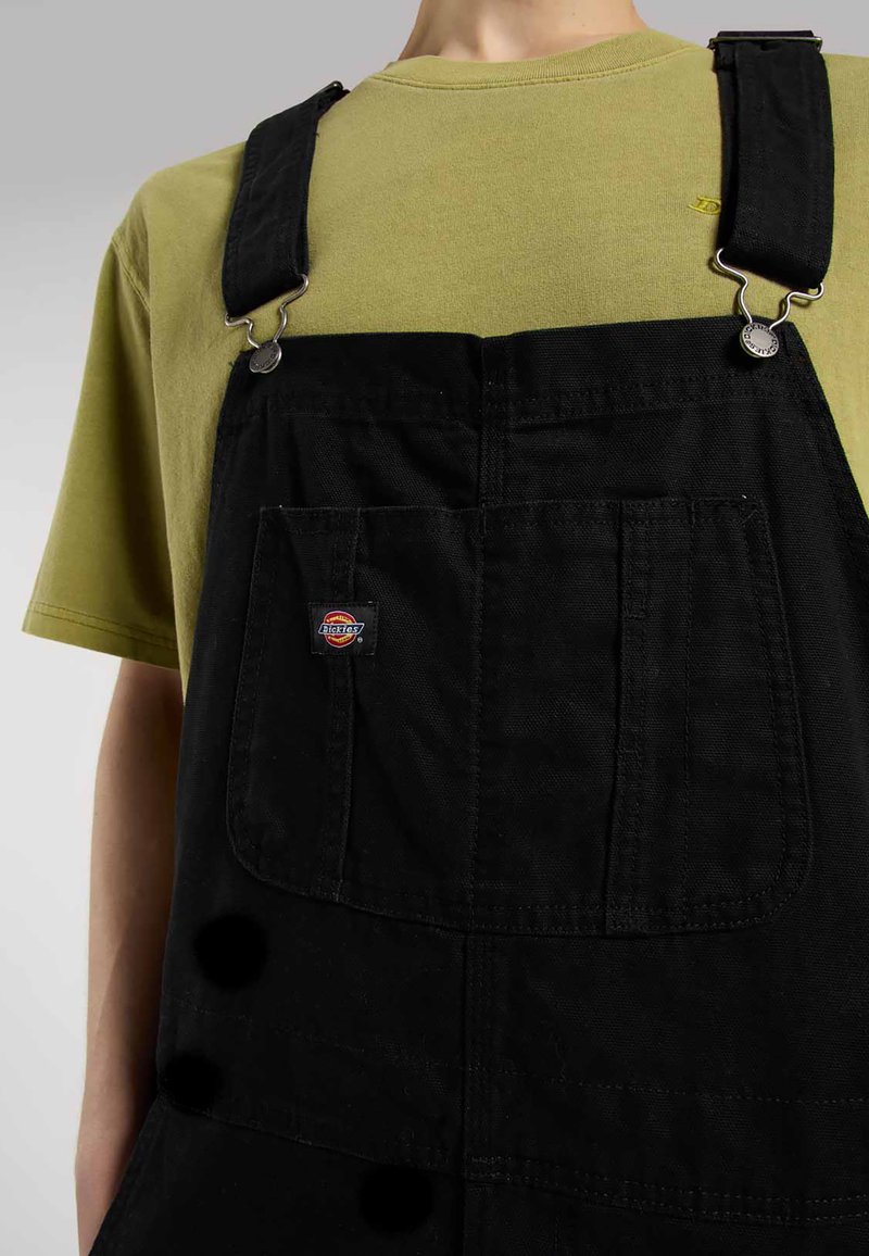 Sorte overalls med justerbare stropper og sølv hardware, med frontlommer og et logo-patch. Olivengrøn t-shirt under.