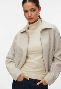 Femme portant un pull à col roulé beige et une veste zippée gris clair, les mains dans les poches de son jean, face à l'avant sur un fond blanc.