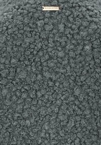 Texturiertes Gewebe in Dunkelgrau mit einer unebenen, lockigen Oberfläche. Enthält ein kleines, metallisches Etikett oben.