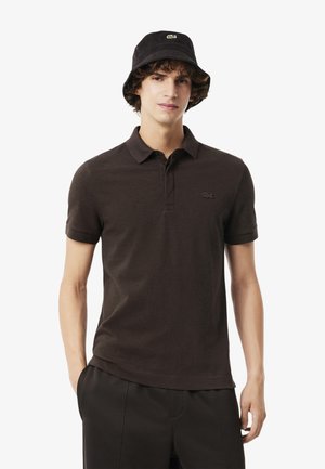 Jonge man in een donkerbruin poloshirt en een bijpassende zwarte bucket hat met een klein groen logo, staande met zijn handen in zijn zakken.