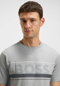 Harmaa puuvillapaita, jossa on pyöreä kaula-aukko, lyhyet hihat ja erottuva 'BOSS'-logo tummemmassa harmaassa. Paidassa on vaakaraitoja valkoisena ja tummana harmaana.