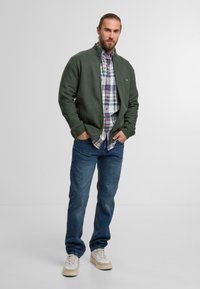 Mann mit Bart in grüner Zip-Jacke, kariertem Hemd, blauen Jeans und weißen Sneakern, der mit den Händen in den Taschen vor einem schlichten Hintergrund steht.