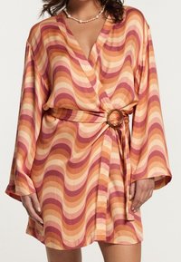 Mujer vestida con un vestido corto tipo envoltura con rayas onduladas de color naranja y rosa, un cinturón con una hebilla redonda y un collar de perlas.