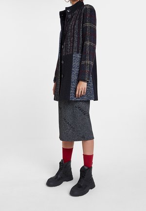 Femme portant un manteau patchwork à carreaux et texturé boutonné, une jupe grise mi-longue, des chaussettes rouge vif et des bottes noires à lacets.