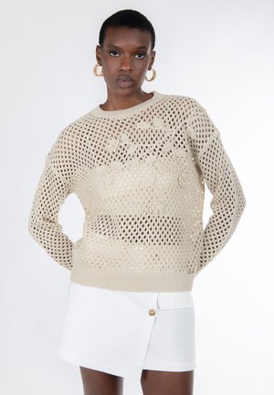 Suéter beige de punto con diseño de malla abierta, con bordado floral en el frente y puños acanalados, combinado con una falda blanca.