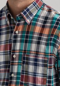 Camisa de botones con un patrón de cuadro multicolor en tonos azul marino, verde azulado, naranja y blanco. Incluye un bolsillo en el pecho y cuello doblado.