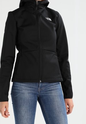 Frau trägt eine enganliegende schwarze North Face Jacke mit Frontreißverschluss und Kapuze, kombiniert mit blauen Jeans vor schlichtem Hintergrund.