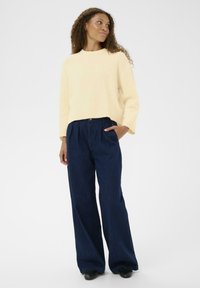 Un pull en tricot côtelé jaune clair avec un col rond, associé à un pantalon en denim foncé à jambes larges et à des chaussures noires, avec une poche latérale sur le pantalon.