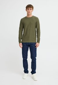 Zelena dolga majica z okroglim izrezom, v kombinaciji z modrimi kavbojkami in belimi supergami. Majica ima strukturirano tkanino in slim fit kroj.