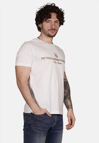 T-shirt bianca a maniche corte con scollo rotondo, con il testo marrone "PATTUGLIA ACROBATICA NAZ." sul davanti e una vestibilità rilassata.