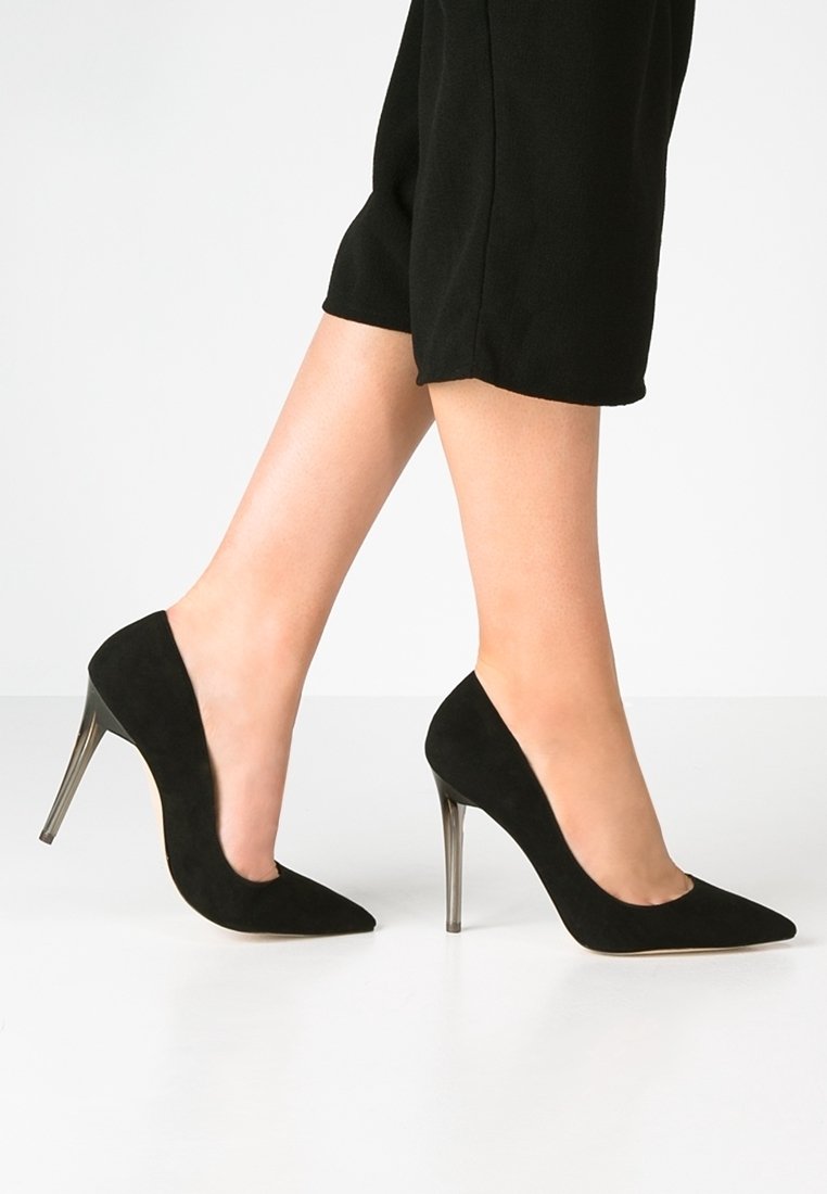 Ben i svarta korta byxor som bär svarta högklackade skor med spetsig tå och metalliska stilettklackar, stående på ett vitt golv.