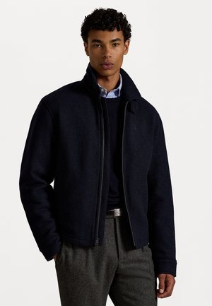 Navy wool-blend jacket with a high collar, zip closure, and ribbed cuffs. Paired with a dark sweater and gray trousers for a layered look.