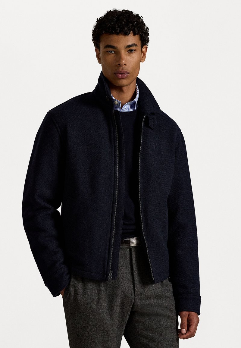 Giacca in misto lana blu navy con colletto alto, chiusura a zip e polsini a costine. Abbinata a un maglione scuro e pantaloni grigi per un look a strati.