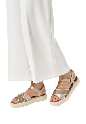Pies con sandalias de plataforma beige con correas de textura tejida y hebillas, combinadas con pantalones anchos blancos.