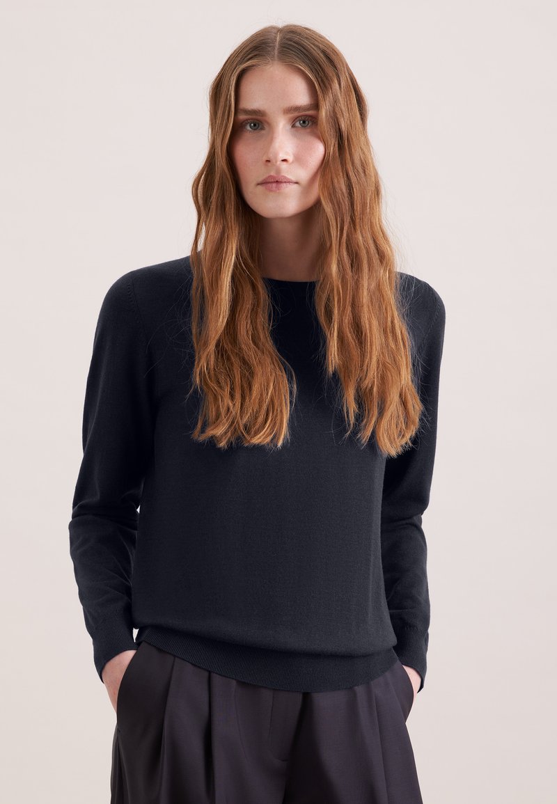 Femme aux longs cheveux ondulés châtain clair, portant un pull noir à manches longues et un pantalon plissé foncé, debout devant un fond uni.