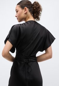 Μαύρο σατέν φόρεμα με ροή στο σχέδιο, κοντά μανίκια και σφιχτή μέση με κόμπο στο πίσω μέρος. Ομαλή υφή και γυαλάδα.