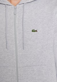Sivá mikina na zips vyrobená z jemného materiálu, s malým zeleným vyšívaným logom krokodíla na hrudníku a nastaviteľnými šnúrkami na kapucni.