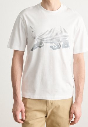 Mand iført en hvid t-shirt med korte ærmer med en stor grafik af en hulkende tyr lavet af små grå former, kombineret med beige bukser.