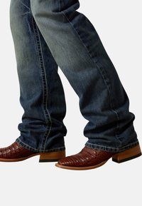 Bruine cowboylaarzen van getextureerd leer, gedragen met donkerblauwe spijkerbroeken, benen gekruist bij de enkels.