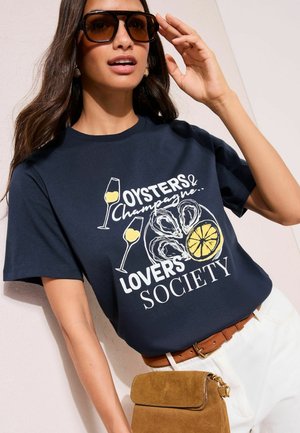 Kvinde iført store brune solbriller, navyblå t-shirt med østers, citron, champagneglas, hvide bukser, brun bælte og holder en tan ruskindtaske.