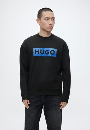 Νέος άνδρας που φοράει μαύρο φούτερ με μπλε ορθογώνιο λογότυπο "HUGO", στέκεται πάνω σε απλό λευκό φόντο.