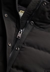 Detalje af sort dunjakke med en glat lynlås, sølvfarvede beslag og syningsaccent. Stoffet har en mat finish med en blød tekstur.