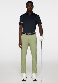 Navy polo shirt with short sleeves, green trousers, black belt, white shoes, and golf club. Smooth fabric with a fitted design and logo details.