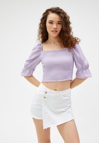 Lavendel crop top met puff-sleeves en smockstructuur, gecombineerd met een witte minirok in wikkelstijl met gouden knopen en een rafelrand.