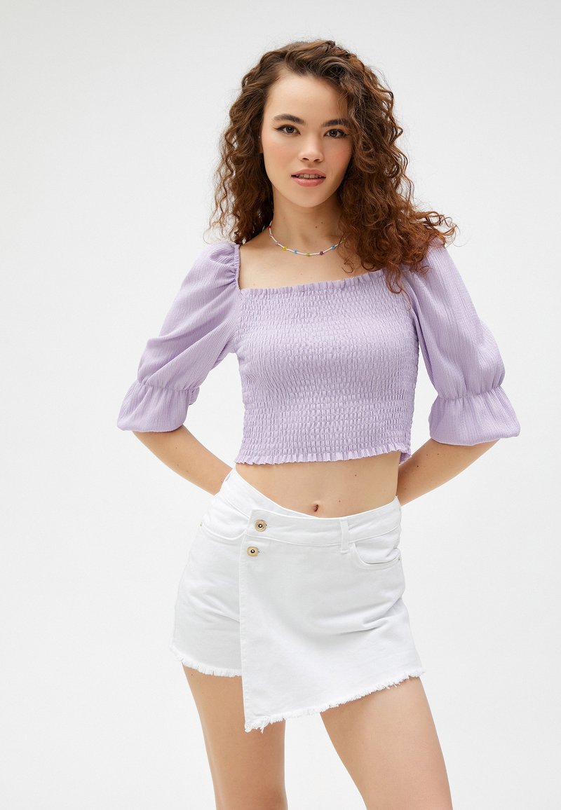 Lavendel crop top met puff-sleeves en smockstructuur, gecombineerd met een witte minirok in wikkelstijl met gouden knopen en een rafelrand.