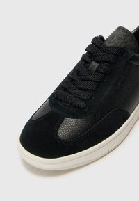 Black leather and suede sneaker featuring textured accents, round laces, and a white rubber sole, with a smooth design and structured shape.
