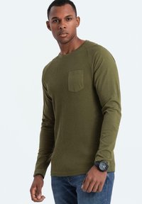 Oliivinvihreä pitkähihainen paita, jossa on ribbitetty tekstuuri, rintatasku ja pyöreä pääntie, yhdistettynä sinisiin farkkuihin ja mustaan kelloon.