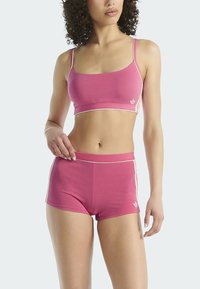 Femme portant un crop top de sport rose et un short assorti avec des bordures blanches et le logo Adidas, se tenant devant un fond uni.