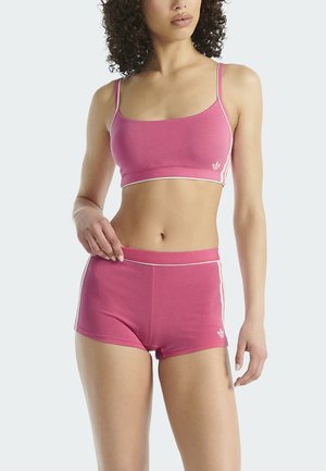 Woman wearing pink athletic crop top and matching shorts with white trim and Adidas logo, standing against plain background.