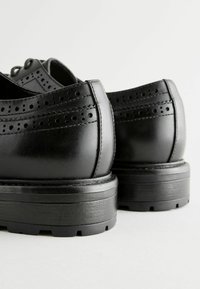 Chaussures en cuir noir avec des perforations, bout arrondi et semelle en caoutchouc noir épaisse. Présentent une finition lisse et des coutures robustes.