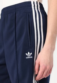 Tamsiai mėlyni sportiniai keliai su baltais šoninėmis juostelėmis, elastinga juosmens juosta ir Adidas logotipu kairėje šlaunies pusėje. Yra šoninės kišenės.