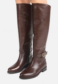 Bottes en cuir marron arrivant au genou avec une finition brillante, fermeture éclair latérale et sangle décorative avec une boucle dorée. Semelle plate, design à embout pointu.