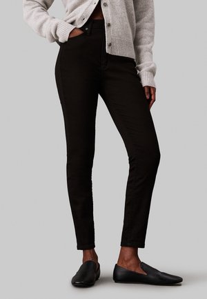 Persona que lleva pantalones ajustados negros, zapatos negros sin cordones y un cárdigan abotonado gris claro, de pie con una mano en el bolsillo.
