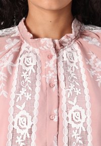 Vaaleanpunainen pusero, jossa valkoiset pitsikoristeet ja kukkakuvioita. Siinä on röyhelökaulus ja napitus edessä.