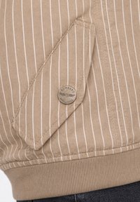 Veste en tissu beige à fines rayures avec une poche à rabat sécurisée par un bouton métallique rond marqué "Bombers Original."